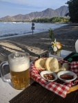 a beer and a plate of food on a table at a restaurant which is on a beach, right next to the sea