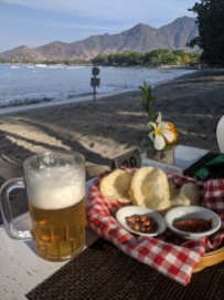 a beer and a plate of food on a table at a restaurant which is on a beach, right next to the sea